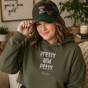 Woman wearing a green hoodie and cap with 'pretty and petty' text in a cozy indoor setting.