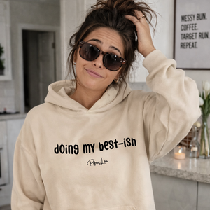 Woman wearing a beige hoodie with text, standing in a kitchen.